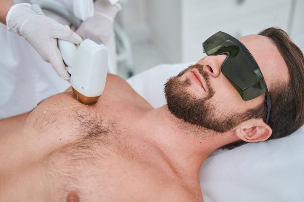 Ein Mann trägt eine Schutzbrille, während eine Fachkraft mit einem Lasergerät eine Enthaarungsbehandlung auf seinem Oberkörper durchführt.